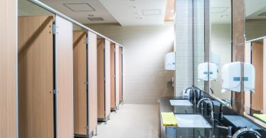 Five restroom stalls line one wall of a public bathroom. On the opposite side are three sinks, soap dispensers, and paper towels.