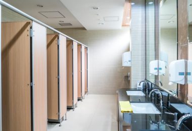 Five restroom stalls line one wall of a public bathroom. On the opposite side are three sinks, soap dispensers, and paper towels.