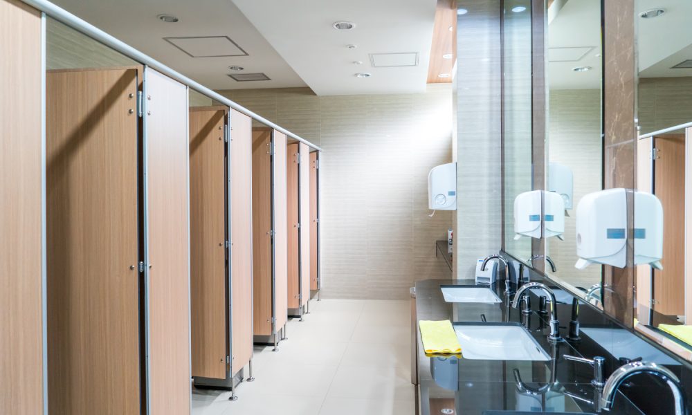 Five restroom stalls line one wall of a public bathroom. On the opposite side are three sinks, soap dispensers, and paper towels.