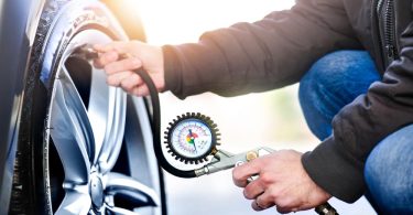 A person wearing a black jacket and jeans checks tire pressure using a gauge connected to the valve stem.