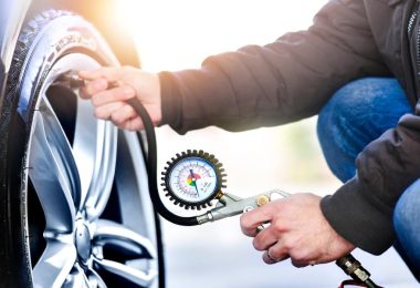 A person wearing a black jacket and jeans checks tire pressure using a gauge connected to the valve stem.