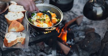A close-up of a person cooking eggs and veggies in a pot above a campfire with bread next to it and a teapot over the fire.