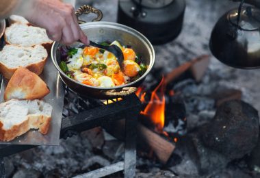 A close-up of a person cooking eggs and veggies in a pot above a campfire with bread next to it and a teapot over the fire.