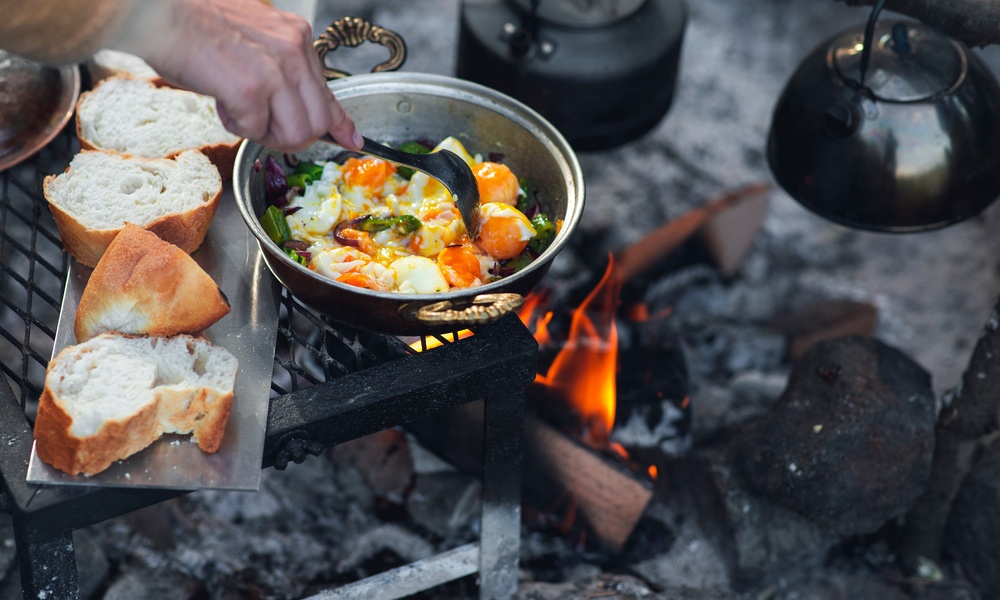 A close-up of a person cooking eggs and veggies in a pot above a campfire with bread next to it and a teapot over the fire.