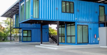 A two-story blue shipping container building with large windows, stacked design, and covered outdoor concrete space.