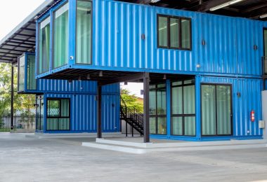 A two-story blue shipping container building with large windows, stacked design, and covered outdoor concrete space.