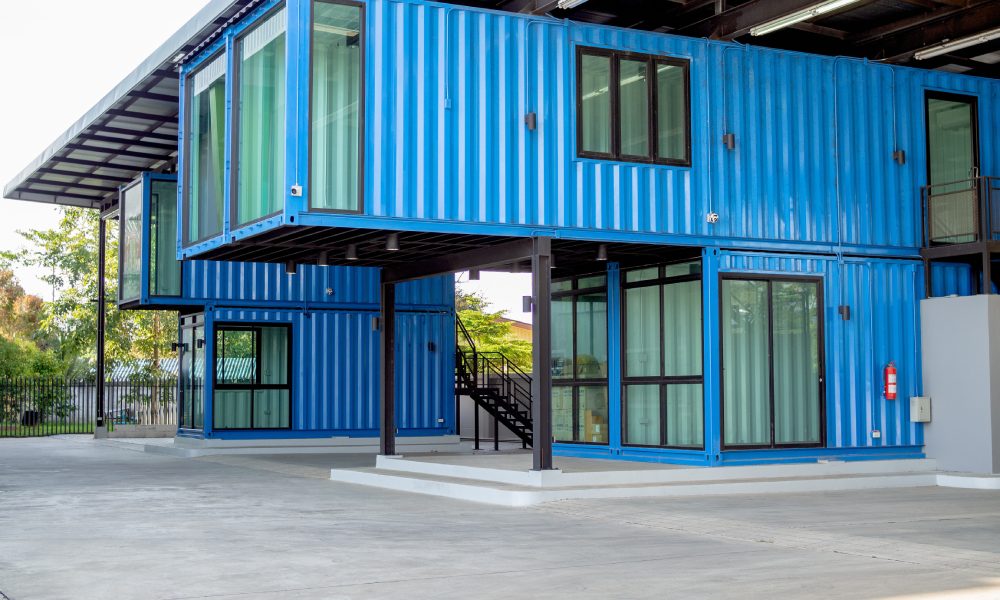 A two-story blue shipping container building with large windows, stacked design, and covered outdoor concrete space.