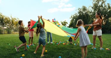 Several kids and two adults standing in a field while using a rainbow parachute to launch colorful plastic balls into the air.