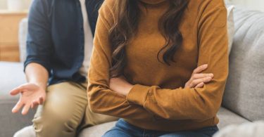A couple on a couch during a disagreement. The woman turns away with folded arms while the man gestures in frustration behind her.
