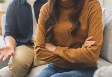 A couple on a couch during a disagreement. The woman turns away with folded arms while the man gestures in frustration behind her.