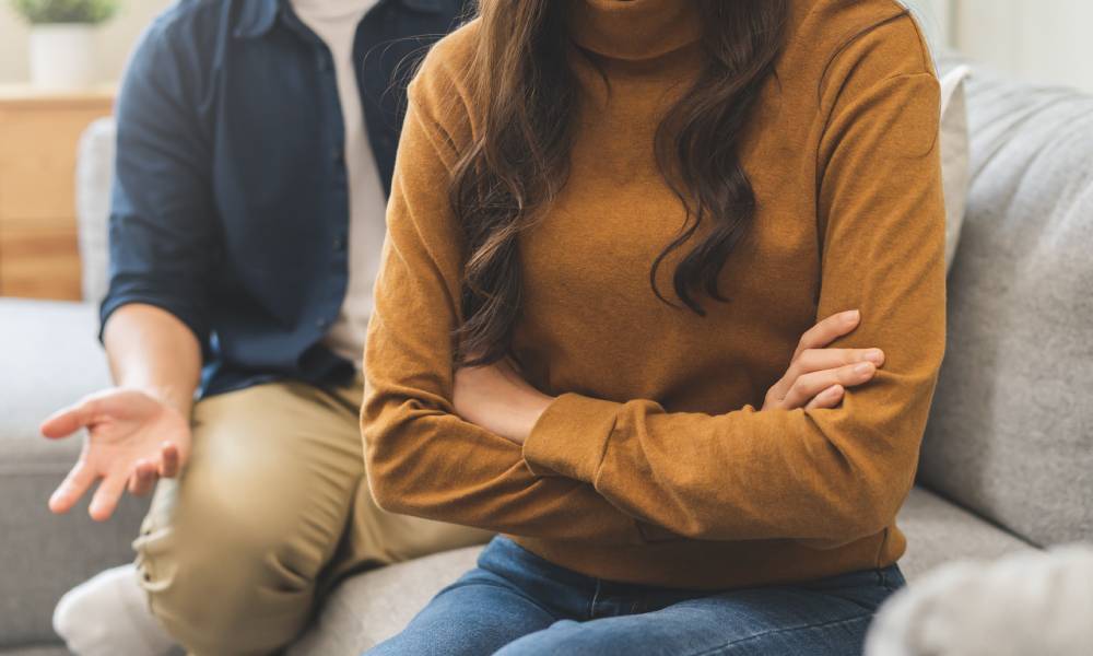 A couple on a couch during a disagreement. The woman turns away with folded arms while the man gestures in frustration behind her.