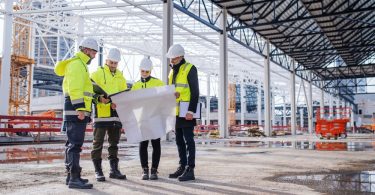 A group of four people in white hard hats and hi-vis jackets standing together at a worksite to look at blueprints.