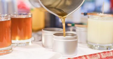 A close-up of someone pouring melted candle wax into small metal tins, with finished candles in glass jars behind.