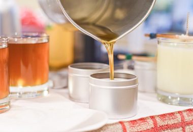 A close-up of someone pouring melted candle wax into small metal tins, with finished candles in glass jars behind.