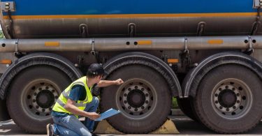 An inspector in a yellow safety vest holds a clipboard, kneels beside a blue tanker truck, and knocks on the middle tire.