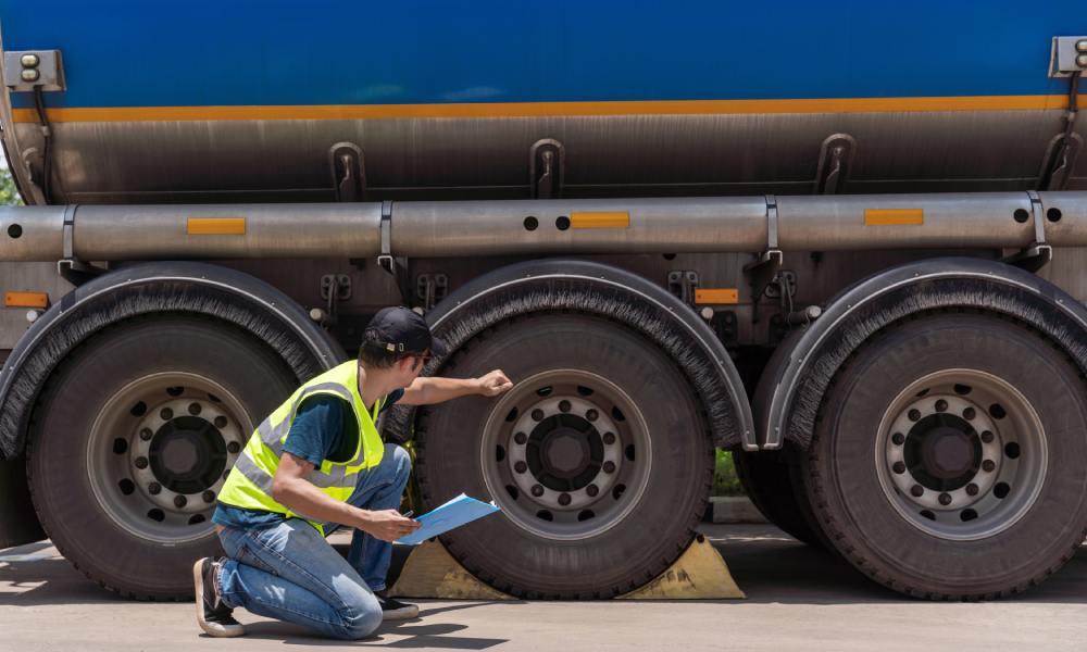An inspector in a yellow safety vest holds a clipboard, kneels beside a blue tanker truck, and knocks on the middle tire.