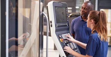 A man and a woman wearing navy blue Polo shirts are looking at the keypad and screen of a CNC machine.