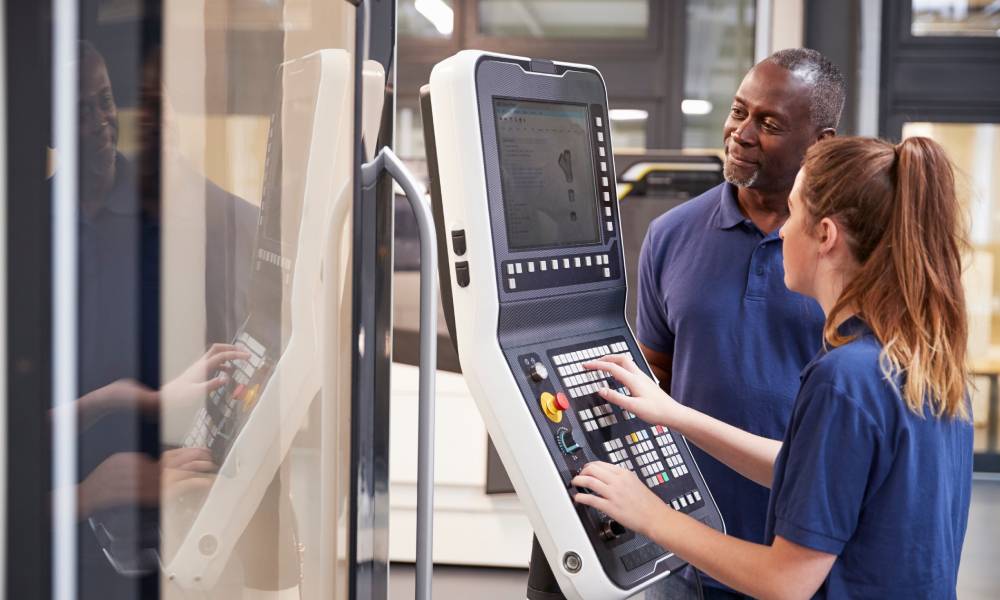 A man and a woman wearing navy blue Polo shirts are looking at the keypad and screen of a CNC machine.