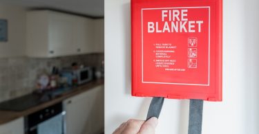 A red “FIRE BLANKET” wall box with instructions, hand-pulling straps, and a kitchen with a stove and cabinets behind.