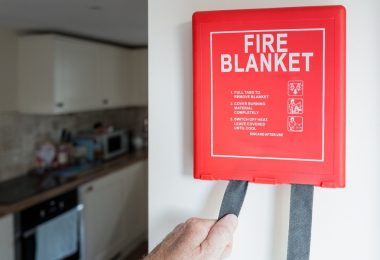 A red “FIRE BLANKET” wall box with instructions, hand-pulling straps, and a kitchen with a stove and cabinets behind.
