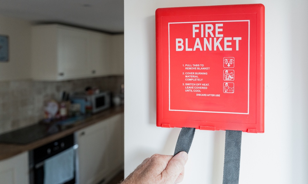 A red “FIRE BLANKET” wall box with instructions, hand-pulling straps, and a kitchen with a stove and cabinets behind.