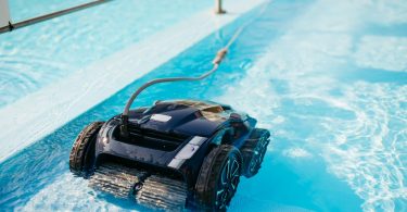 A black robotic pool cleaner with wide rubber wheels actively scrubs the floor of a sunlit blue swimming pool.