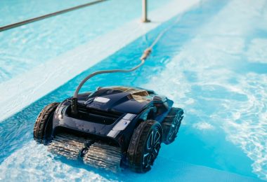 A black robotic pool cleaner with wide rubber wheels actively scrubs the floor of a sunlit blue swimming pool.