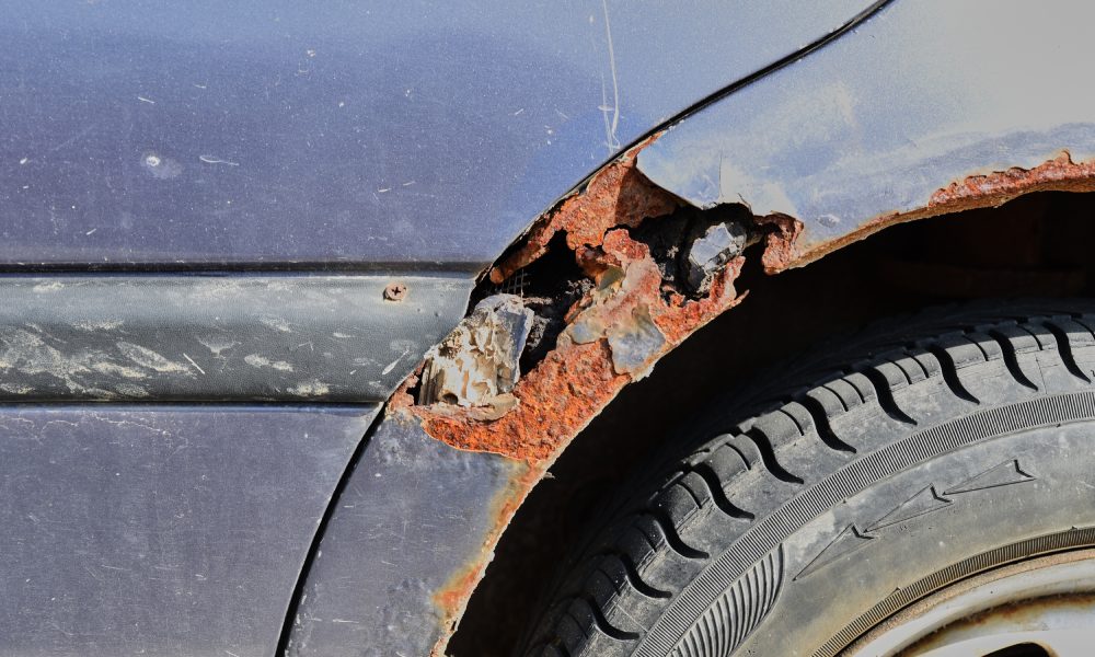 Sunlight shining down onto a rusty blue car parked outside. Rust causes the wheel well to turn orange and break apart.