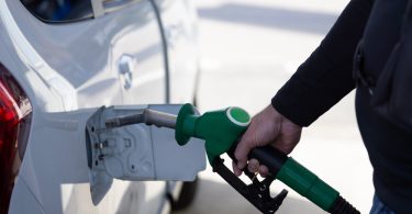 A close-up of a parked white SUV. Someone wearing a black shirt holds a green fuel nozzle up to the car's gas tank.