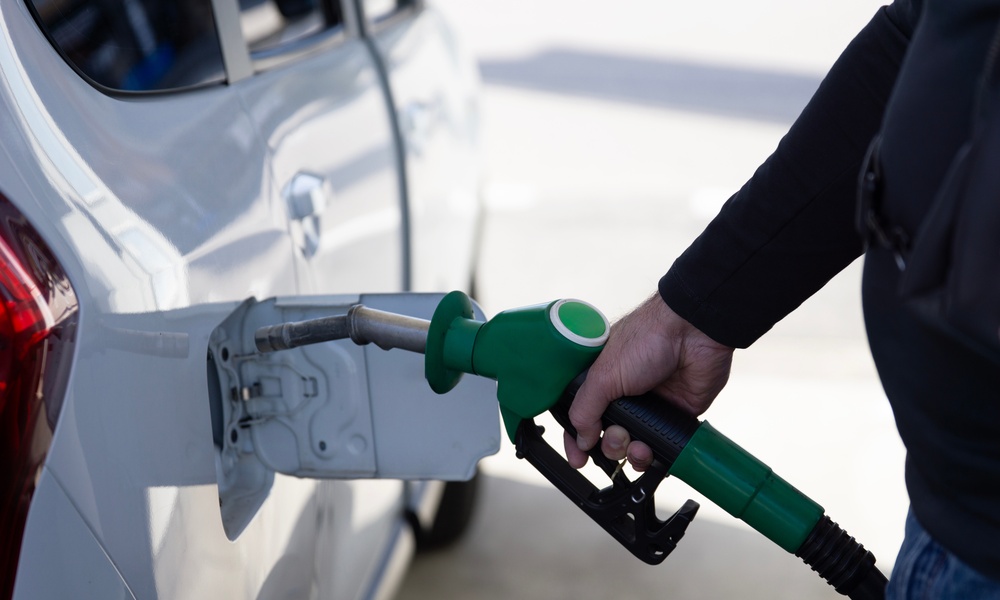 A close-up of a parked white SUV. Someone wearing a black shirt holds a green fuel nozzle up to the car's gas tank.