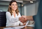 A woman wearing a white blouse smiles as she shakes hands with someone across a table containing documents.