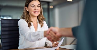 A woman wearing a white blouse smiles as she shakes hands with someone across a table containing documents.