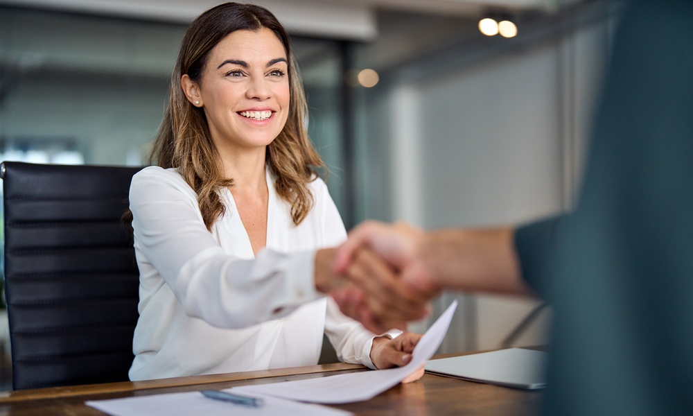 A woman wearing a white blouse smiles as she shakes hands with someone across a table containing documents.