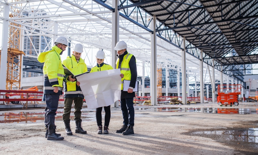 Four construction professionals in safety vests looking at the blueprints of a building that is under construction.