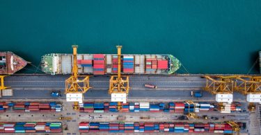 A shipping container ship is docked at port. The ship is already loaded with containers but is being loaded with more.