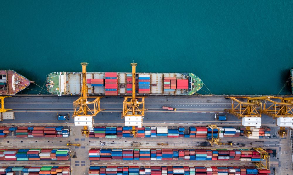 A shipping container ship is docked at port. The ship is already loaded with containers but is being loaded with more.