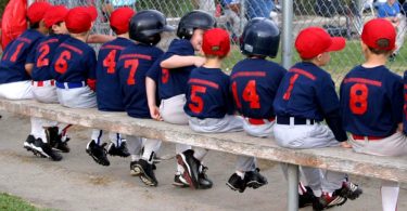 A little league baseball team is sitting on a long bench. The kids are all wearing the same uniform.