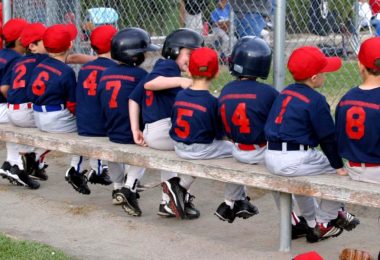 A little league baseball team is sitting on a long bench. The kids are all wearing the same uniform.