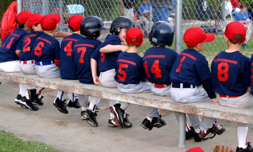 A little league baseball team is sitting on a long bench. The kids are all wearing the same uniform.