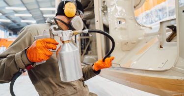 A worker in protective gear sprays a coating onto large metal machinery inside an industrial workshop.