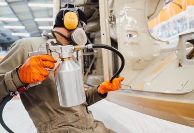 A worker in protective gear sprays a coating onto large metal machinery inside an industrial workshop.