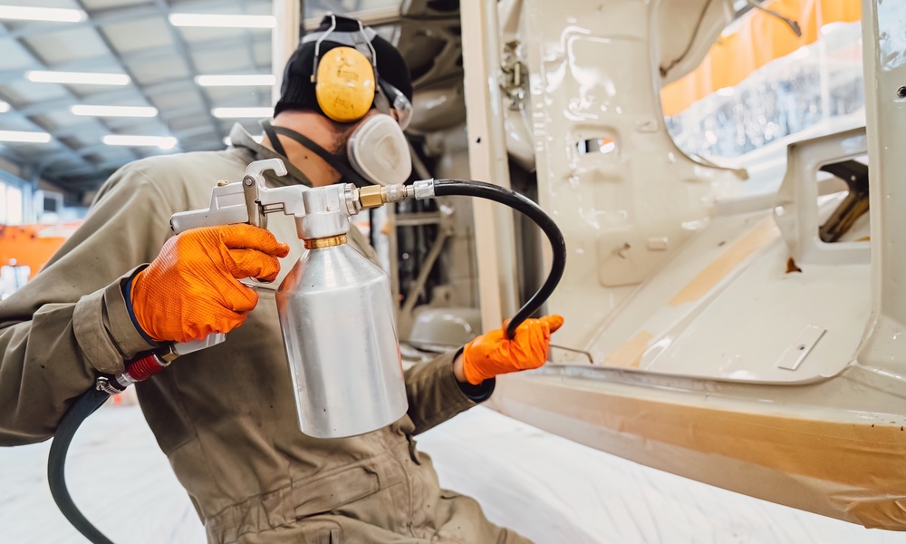 A worker in protective gear sprays a coating onto large metal machinery inside an industrial workshop.