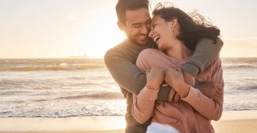 A smiling couple embraces on a sandy beach at sunset, laughing together as ocean waves roll in behind them.