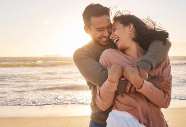 A smiling couple embraces on a sandy beach at sunset, laughing together as ocean waves roll in behind them.
