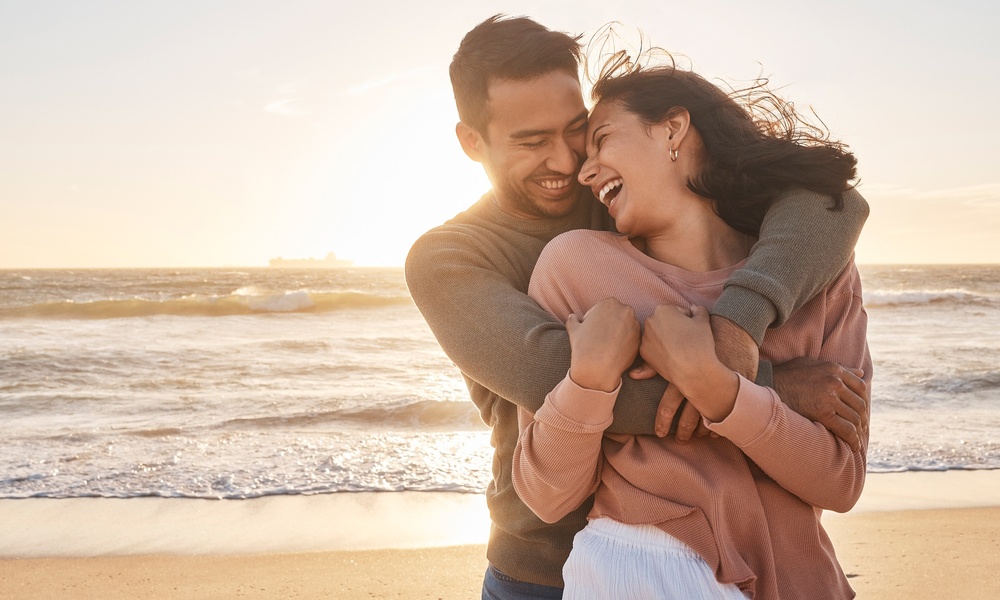 A smiling couple embraces on a sandy beach at sunset, laughing together as ocean waves roll in behind them.