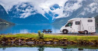 An RV is parked next to a lake and a picnic table. Far in the background is a clouded mountain range.