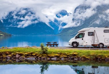 An RV is parked next to a lake and a picnic table. Far in the background is a clouded mountain range.