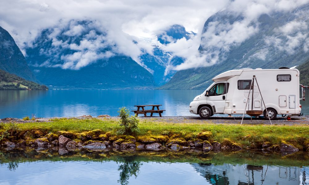 An RV is parked next to a lake and a picnic table. Far in the background is a clouded mountain range.