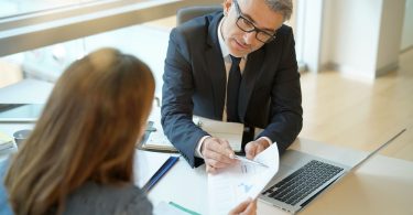 A man wearing a suit and tie sits at a table across from another person. He is pointing to a document.