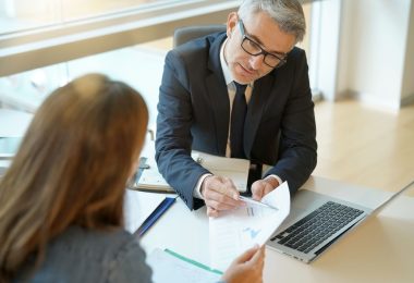 A man wearing a suit and tie sits at a table across from another person. He is pointing to a document.